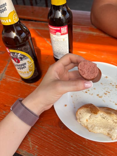 The image shows a close-up view of a person's hand holding a sliced sausage over a white plate with a piece of bread on it. The sausage appears to be a raw, pinkish-brown color with visible pores. The bread is crusty on the outside and soft and white on the inside. In the background, there are two bottles of beer on an orange wooden table. The label on the left bottle is partially visible and reads "Schlaffe Seppel Adler." The table is outdoors, likely at a beer garden or outdoor seating area. A portion of a person's leg is visible in the upper right corner. This scene could be from Obernburg am Main, Germany, given the context of German beer and sausages.
