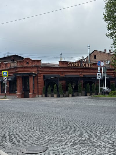 The image shows the "Syndicate Cafe" building in Akhieli, Georgia. It is a brick building with awnings over the windows and decorative plants along the front. A cobblestone street runs alongside the building. Several traffic signs are visible, including a pedestrian crossing sign and a no-trucks sign. The sky is overcast. Overhead electrical wires cross the frame. Part of a tree is visible in the top left corner.