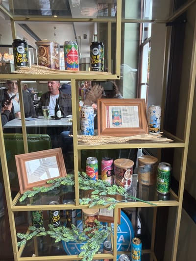 An indoor scene displays a gold-framed shelving unit filled with various items, including canned beverages, jars, and decorative elements. The shelving unit is positioned in what appears to be a public establishment, possibly a bar or restaurant, with people visible in the background.

The top shelf features two cans of "Son of a Beach" beer, a jar filled with dried citrus slices and other items, and a can labeled "JUDECIOUS JUICY PALE." Several stalks of wheat are arranged in front of these items.

The second shelf contains a framed picture with text and an illustration of a beer can, a tall can with a blue and yellow design and "NEW YORK TRAVELLING TAXI" text, and a can with a red and green design. Decorative green hop sprigs are placed in front of the framed picture and the cans.

The third shelf has two small jars with wooden lids, one containing dried fruit slices and the other a beige substance, alongside a can of "OAKHAM CITRA SESSION IPA." More green hop sprigs are displayed here.

The bottom shelf shows several more cans of beer, including "Son of a Beach" and cans with green and black designs. A large blue circular object with white lettering is also visible. Additional green hop sprigs are present.

The overall lighting suggests an indoor environment with ambient light, possibly from windows or overhead fixtures. The establishment appears to be busy, with patrons seated at tables in the background and staff members attending to them. The location is identified as being in Epsom and Ewell, United Kingdom.