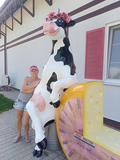 A woman wearing a pink baseball cap, a grey tank top, and denim shorts stands leaning against a large, black and white cow statue adorned with a floral headdress. The cow statue is positioned next to a sculpture of a wedge of cheese, which appears to be a bench. The woman has her arms crossed and is looking at the camera with a slight smile. The scene is outdoors, with a paved walkway, green grass, and a white building with brown trim and red shutters in the background. The location is identified as Shabo, Ukraine. The lighting suggests it is daytime, with bright sunlight casting shadows.