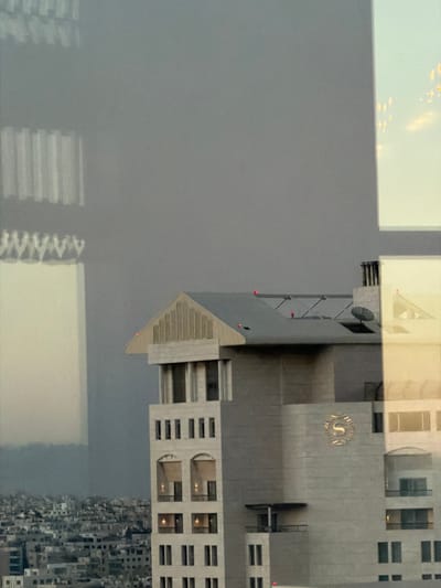 This is a daytime, medium-angle, eye-level outdoor shot of a modern building in Amman, Jordan, viewed through a window with reflections. The primary subject is a light-colored, multi-story building with a prominent "S" logo on its facade, which appears to be the Sheraton Hotel. The building has several floors with distinct window arrangements, including arched openings and rectangular windows. Red lights are visible on the rooftops of this building and another adjacent structure, indicating potential navigational lights or markers.
To the left and background, a densely populated urban landscape is visible, characterized by numerous smaller buildings and structures. A large, somewhat indistinct building or structure with vertical lines is reflected or appears in the foreground on the left, possibly another building or a distorted reflection from the window. The sky is overcast and gray, suggesting either a cloudy day or haze, with some subtle warm light in the upper right quadrant, hinting at the sun's position. The overall impression is one of an urban environment, likely viewed from an elevated position such as another building or hotel room.