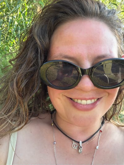 A medium-close-up selfie taken outdoors shows a smiling woman wearing large black oval sunglasses and a light-colored tank top. Her brown, wavy hair falls around her face and shoulders. She is wearing two necklaces: one is a black cord with a star charm and a shoe charm, and the other is a silver chain with several small gems. The background consists of blurred green foliage, suggesting an outdoor setting like a park or garden in Kamianka-Buzka, Ukraine. The lighting indicates it is daytime.