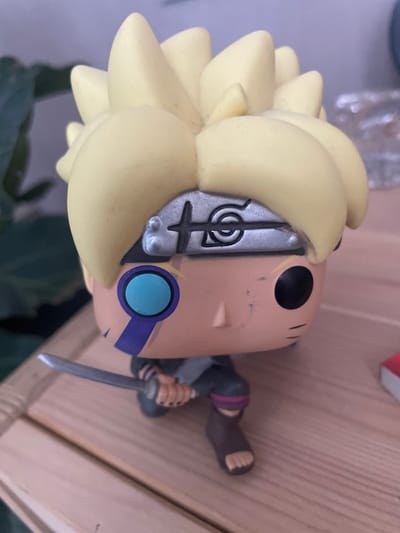 A close-up, eye-level shot shows a Boruto Uzumaki Funko Pop figurine positioned on a wooden surface. The figurine has bright yellow spiky hair, a silver headband with the Konoha village symbol, and a teal circular mark on its cheek with a purple stripe beneath it. It holds a silver katana in its right hand and wears a dark outfit with pink accents around its knees and brown footwear. The background is softly blurred, with a green plant on the left and what appears to be a clear glass object on the right. The wooden surface has visible grain and is lit from above, casting subtle shadows. There is no indication of the city of Rosinka, Russia, in this image.