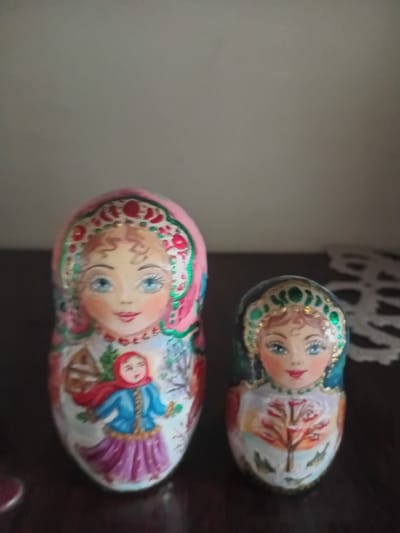 The image displays two traditional Russian nesting dolls, also known as Matryoshka dolls, positioned on a dark, reflective surface. The doll on the left is larger and predominantly pink, adorned with a detailed floral headdress and a painted portrait of a woman with blue eyes and reddish-blonde hair. This doll also features a smaller doll painted on its midsection, depicting a child in a blue jacket and red scarf. The doll on the right is smaller and has a dark teal headdress with gold accents. It also shows a painted portrait of a woman with blue eyes and similar reddish-blonde hair. The background is a plain, neutral color, with a white lacy doily-like object visible in the upper right corner of the image. There is no discernible connection to the city of Kherson, Ukraine.