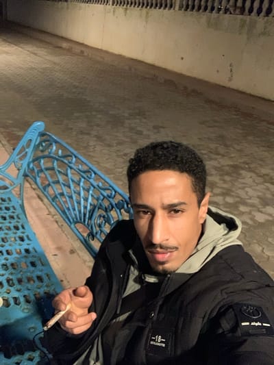 A man with dark curly hair and a slight mustache poses in a dark setting. He wears a black jacket over a hooded sweatshirt and holds a lit cigarette or joint. He sits on a bright blue metal bench with an intricate design. A concrete wall and what appears to be a drainage system are in the background, adding to the urban and somewhat gritty atmosphere.