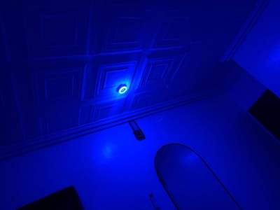 The image shows an interior space illuminated with blue light. The ceiling has a grid-like pattern with a circular light fixture at the center. A wall-mounted object is visible below the ceiling, and a mirror is positioned on the lower right side of the wall. The scene is in Rumuigbo, Nigeria.