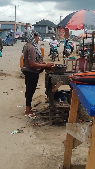 A woman with braided hair is standing in a street market in Warri, Nigeria. She is wearing a brown t-shirt and black leggings, and she is interacting with a bowl of food or goods on a wooden stand. In the background, there are several motorcycles, people on motorcycles, buildings, and street vendors with umbrellas and stalls. The ground is unpaved and littered with debris. The sky is overcast.