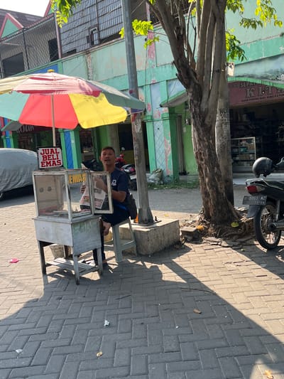 The image depicts a street scene in Juwet Kenongo, Indonesia, on a sunny day. In the foreground, a man sits on a stool next to a mobile cart, which appears to be a jewelry or pawn shop stall. A sign above the cart reads "JUAL BELI EMAS," meaning "Buy Sell Gold." The cart has a glass display case, and the man is holding what looks like a newspaper or brochure. The cart is positioned on a paved sidewalk, with a tree to the right. A motorcycle is parked behind the tree, and a parked car is partially visible to the left. The background shows buildings with green and pastel facades, typical of Indonesian architecture, and foliage from trees. The overall impression is of a bustling local marketplace or commercial street.