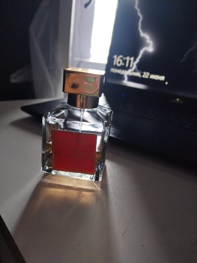 A perfume bottle, identified as Baccarat Rouge 540, rests on a surface, casting a shadow. In the background, a laptop displays the time as 16:11 and the date "понедельник, 22 июня", suggesting it is Monday, June 22nd. The laptop screen also features an image of a lightning strike. The overall environment appears to be indoors, with ambient lighting. There are no people present in the image. The location is not specified to be Chita, Russia.