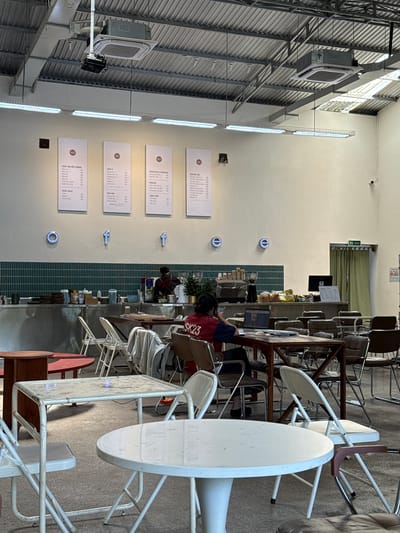 The image depicts the interior of a modern coffee shop in Tam Kỳ, Vietnam, with a rustic industrial aesthetic. The space features high ceilings with exposed ductwork and lighting fixtures. The walls are painted white, with a green tiled backsplash behind the counter. Four large menu boards are mounted on the wall, displaying drink options. The words "o f f e e" are spelled out in neon blue lights across the lower portion of the wall.
In the foreground, several white metal folding tables and chairs are arranged, some with worn surfaces. Further back, a more substantial wooden table is occupied by a person wearing a red and blue shirt with "SK23" visible, working on a laptop. This individual is seated facing away from the camera, towards a counter area where a barista is partially visible.
The counter area is equipped with a coffee machine, stacks of cups, and various other items associated with a coffee shop. Bags of bread and other food items are also displayed on shelves behind the counter. Various seating arrangements, including cushioned chairs and more metal chairs, are scattered throughout the space. A green curtain partially obscures a doorway labeled "TOILET" in the background. The lighting suggests it is daytime, with natural light filtering in from unseen sources. The overall atmosphere is casual and contemporary.