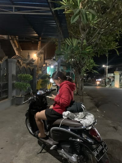 A young woman in a red hoodie and black shorts is sitting on a parked black scooter at night on a street in Buleleng, Indonesia. She is looking down at her phone, which she is holding in her hands. The scooter is parked on the side of the road, which is paved and has streetlights. There are buildings and trees on either side of the road. In the background, there is a gate with decorative elements and potted plants. A stack of clothing is on the back of the scooter behind the woman. The license plate of the scooter reads "DK 6966 VV 09-26".