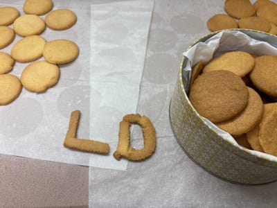 Several round, tan-colored cookies are on a countertop, some on parchment paper and some inside a round tin with a white and gold patterned exterior. On the parchment paper, the letters "LD" are constructed from cookie dough. A group of additional round cookies are arranged on the paper, adjacent to the letters. The countertop is a light gray color.
