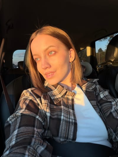 A woman is seated in a car wearing a white t-shirt and a brown and tan plaid flannel shirt. She is wearing a seatbelt. She has blonde hair and a gold geometric earring in her left ear. The scene is illuminated by warm, golden sunlight. The location is unclear, and there is no indication of being in Rogachovo, Russia.