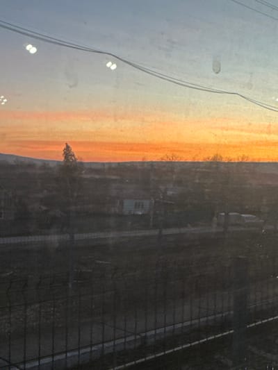 The image depicts an outdoor landscape scene at either sunrise or sunset, viewed through a dirty or reflective window, originating from Ungheni, Romania. The sky occupies the upper two-thirds of the frame, displaying a gradient from a vibrant orange-red at the horizon to a pale blue at the top. Multiple circular light reflections and smudges are visible on the glass surface in the upper portion. Two parallel, dark utility wires or cables traverse the upper sky diagonally from left to right.
The mid-ground reveals a semi-rural or low-density urban environment under low light conditions. The terrain is largely flat, with distant hills visible on the far left horizon. Sparse, deciduous trees and shrubs are scattered across the landscape, appearing bare, suggesting a colder season. A prominent, solitary conifer-like tree stands silhouetted against the bright orange sky on the left side of the mid-ground. Several indistinct residential buildings or houses are discernible, with one light-colored structure particularly visible to the right of the prominent tree. A horizontal band of darker ground, possibly a road or cleared area, stretches across the mid-ground. On the far right, faint outlines suggest the presence of one or more vehicles, possibly a white van.
The immediate foreground is dominated by a dark metal fence, consisting of vertical bars and horizontal supports, running across the lower third of the image, obscuring the ground directly beneath it. No people or discernible actions are present in the scene.