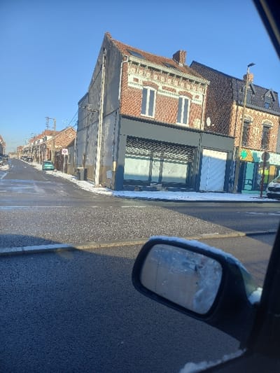 The image displays a street intersection in Noyelles-sous-Lens, France, under a clear blue sky, indicating daylight. Patches of melting snow and ice are visible on the ground, rooftops, and along the road edges, suggesting cold weather or winter conditions. The perspective is from inside a vehicle, with the passenger-side rearview mirror, partially obscured by snow or ice, prominent in the lower right foreground.
In the center-right, a two-story brick building with a sloped terra-cotta tiled roof stands at the corner. Its upper level features two tall rectangular windows and decorative white brickwork. The ground floor presents a dark grey facade with a partially lowered metal security shutter covering large display windows, suggesting a closed commercial establishment. Immediately adjacent to its right is another multi-story brick building with a dark mansard roof and dormer windows. Its ground level features a storefront with a visible green sign fragment indicating "PHARMACIE PRIX," identifying it as a pharmacy. To the left of the central corner building, a plainer structure with a light-colored, possibly stuccoed, facade and a sloped tiled roof faces the intersection.
A street extends to the left, lined with a row of similar residential or commercial buildings receding into the distance. A light blue compact car is parked on this street further down. Asphalt roads are visible, appearing wet in places, with textured paving at the immediate intersection. A white vehicle is partially visible on the far right, in front of the pharmacy. Utility lines are strung between poles or attached to buildings along the left street, which also features streetlights. No individuals are visible in the scene.