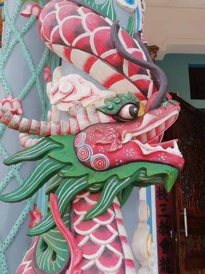 The image shows a close-up view of a colorful, decorative dragon sculpture on a pillar. The dragon is primarily painted in shades of red, pink, green, and white, with intricate details such as scales, eyes, and fangs. The dragon's head is prominently featured, with its mouth open, revealing its teeth and tongue. The pillar itself appears to be part of a larger structure, possibly a temple or shrine, with elements of traditional Asian architecture visible in the background, including a wooden door with carvings and a blue wall with a lattice pattern. There are no people or animals in the image. The location is identified as Lộc Quý, Vietnam.