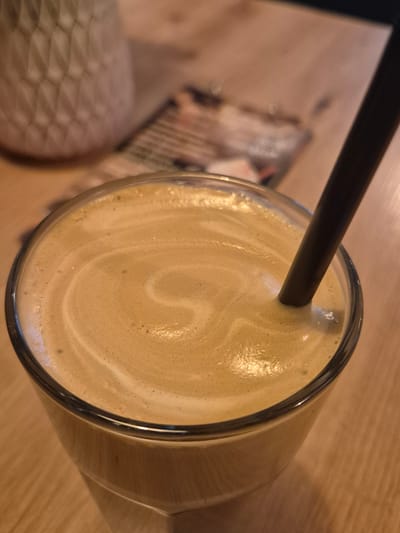 The image shows a top-down view of a beverage in a clear glass, likely coffee or a similar drink with a frothy, light brown surface exhibiting swirling patterns. A black straw is inserted into the liquid. The glass rests on a light-colored wooden table. In the blurry background, there's a patterned chair and a menu. No people are visible. The location, Pavlikeni, Bulgaria, cannot be determined from the visual data alone.