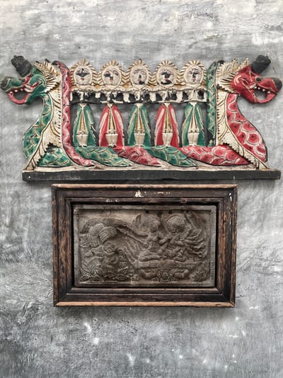The image shows two pieces of art displayed on a textured gray concrete wall. The upper artwork is a colorful wooden carving featuring two dragon heads on either side, with a row of five figures with distinct facial features and elaborate attire in between. The figures appear to be standing with their arms held down, and their clothing is depicted in red and green with white accents. The lower artwork is a framed stone relief carving. The frame is made of dark, weathered wood. The relief depicts a scene with multiple figures in a stylized, detailed manner, suggesting a narrative or mythological subject. The overall aesthetic suggests traditional Indonesian folk art or cultural artifacts. The location is identified as Karangkebagusan, Indonesia, indicating a specific regional context for these art pieces.