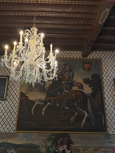 The image captures a room in what appears to be a historic building, potentially in Bunyola, Spain. Dominating the view is an ornate, multi-tiered crystal chandelier. The ceiling is decorated with dark wood panels accented with light-colored decorative strips. A large painting hangs on the wall, depicting a man on horseback in what seems to be period attire, complete with armor and a hat. The painting also features a coat of arms and some text in a foreign language, potentially Spanish. The backdrop in the painting shows a landscape and what looks like a city or structure. The wallpaper behind the painting has a geometric pattern. The presence of the painting, chandelier, and the decor suggest that this is likely a room within a historical building or museum. The overall lighting seems subdued, with the chandelier providing the main source of light. The composition includes elements that are typical of European historic settings, such as the painting and the elaborate chandelier.