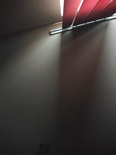 The image shows a dimly lit interior space, possibly a room in a residential building in Kladno, Czechia. A portion of the ceiling is visible at the top, with red blinds casting shadows on a dark wall. A white air conditioning unit is mounted on the wall in the upper right quadrant. In the lower right corner, a small, dark object, possibly a decorative item or a piece of hardware, is visible on the floor. The overall lighting suggests low ambient light with a directed light source creating the shadows from the blinds.