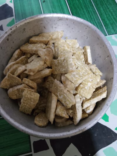 In Merbopuntong, Indonesia, a close-up, overhead shot shows a metal bowl filled with sliced tempeh. The tempeh is cut into irregular, bite-sized pieces and appears to be raw or unseasoned. The bowl is placed on a surface with a geometric pattern of green, white, and black. The lighting is dim, casting soft shadows.