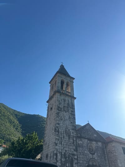 This image depicts the Church of the Nativity of the Blessed Virgin Mary in Prčanj, Montenegro, under a clear blue sky on a sunny day. The central subject is the church's prominent bell tower, constructed from weathered grey stone blocks. The tower is square-shaped, featuring arched belfry openings near its top and a pyramidal dark-colored roof capped with a cross. Below the belfry, a circular indentation is visible on the stone facade. To the right of the bell tower, a portion of the main church building is visible, also constructed from similar stone blocks. This section displays a gabled roof with a cross at its peak and two circular windows, one larger (resembling a rose window) below the gable and a smaller one above it. An adjacent stone building with a terracotta tiled roof is partially visible to the far right. In the background, lush, densely forested green mountains rise under a brilliant, cloudless blue sky. A strong lens flare or sun glare is evident in the upper right quadrant of the sky. The very bottom of the frame shows a dark-colored vehicle. No individuals are present in the scene.