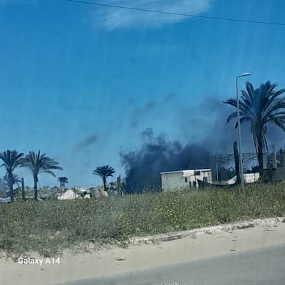 The image depicts a scene in Nuseirat, Palestinian Territory, featuring several palm trees lining a street. A plume of black smoke rises into the bright blue sky behind buildings adjacent to the street. The scene appears to be taken from a moving vehicle, as evidenced by the "Galaxy A14" watermark, presumably the camera used. The street is bordered by vegetation. A streetlight is visible among the palm trees.