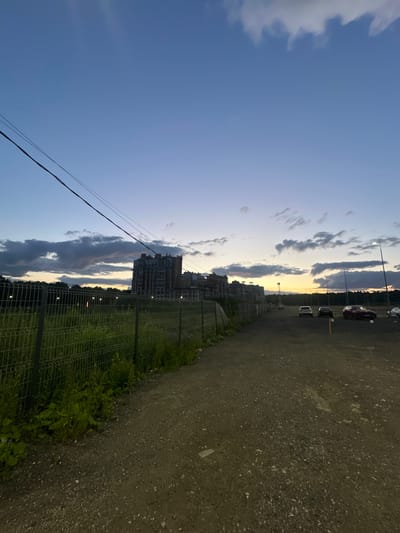 The image shows a desolate, unpaved road leading into the distance under a twilight sky. Tall, multi-story residential buildings are visible in the background to the left, partially obscured by a wire fence and overgrown vegetation. Several cars are parked on the right side of the road. The sky displays hues of deep blue transitioning to orange and yellow near the horizon, with scattered dark clouds. Power lines stretch diagonally across the upper portion of the frame. The scene appears to be on the outskirts of Laikovo, Russia, likely a developing or industrial area given the unfinished road and presence of construction. No people are visible in the image.