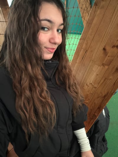 A young female with long, wavy brown hair and dark eyes is depicted from the upper torso up, looking directly at the camera with a slight smile. She wears a black puffer jacket or vest over a dark long-sleeved garment, with a light-colored, ruched sleeve covering her left forearm. The background features a diagonal light-brown wooden beam or panel on the right, behind which a green mesh net is visible. Below the wooden element, a bright green surface, possibly artificial turf, is present, on which a dark garment with a partial white logo (resembling "The North Face") rests. The scene is located in Souffelweyersheim, France, likely within an indoor recreational or sports facility.