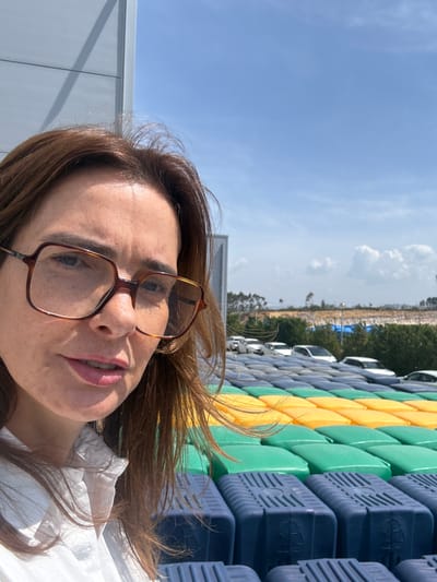 A woman with brown hair and brown-framed glasses is featured prominently in a selfie-style photograph, looking directly at the camera with a neutral to slightly open-mouthed expression. She is wearing a white collared shirt. The image is taken outdoors on a bright, sunny day with a clear blue sky dotted with scattered white clouds.
In the background, a large, colorful structure is visible, composed of numerous interlocking modular blocks in alternating rows of yellow, green, and dark blue. On the dark blue blocks, a circular symbol resembling a recycling logo or a stylized 'R' is discernible. This structure could be part of a floating dock system or a temporary event setup. Beyond these modules, many white cars are parked in orderly rows, indicating a large parking lot, possibly for a commercial facility or an event. Further in the distance, a dry, open landscape with sparse vegetation and hills stretches under the bright sky. The strong sunlight casts clear shadows, highlighting the outdoor setting.
The location is an open-air environment, possibly near a coastal or industrial area in Torres Vedras, Portugal, given the general landscape and the type of structures present. There is no other distinct text visible apart from the symbols on the blue blocks.