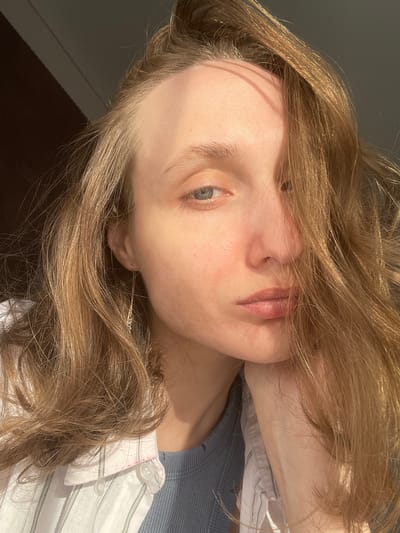 A close-up, slightly upward-angled portrait shows a young woman with light blonde hair, likely indoors. Her hair is long and wavy, partially obscuring her face and shoulders. The woman has pale skin, light blue eyes, and subtle features. She is wearing a light blue t-shirt under a white button-down shirt with thin grey stripes. Part of her right hand is visible near her neck. The background is a dark brown, possibly a wall or furniture. The lighting is bright, casting strong shadows on her face and hair. There's no information in the image to indicate that the picture was taken in Sepych, Russia.