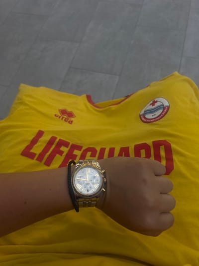 The image shows a person's arm wearing a gold Michael Kors watch and a black braided bracelet, resting on a yellow t-shirt. The t-shirt has red lettering that says "LIFEGUARD" and a circular emblem with a red cross and Cyrillic text that translates to "Water Rescue Service". The t-shirt appears to be part of a uniform for lifeguards in Banya, Bulgaria. The background is a grey tiled floor.