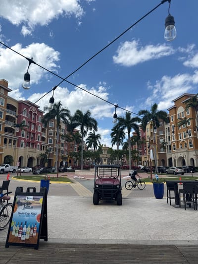 The image depicts a street scene in Naples, Florida, USA, under a partly cloudy sky. Multi-story buildings with diverse architectural styles and colors line one side of the street, interspersed with palm trees. Parked cars are visible along the building side of the street. Overhead, string lights with exposed bulbs are strung across the street.
On the right side of the frame, near the bottom, is a sidewalk with a wooden plank edge. A sandwich board sign advertising alcoholic beverages is placed on the sidewalk. Further along the sidewalk, there is a red utility vehicle with an open back and a bicycle ridden by a person with a backpack. There are tables and chairs visible in the background. A large blue planter sits by the side walk.