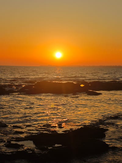 A vertical, low-angle shot depicts a sunset over the sea in Costa Oeste, Spain. The sun, a bright white orb, is positioned high in the orange sky, casting a golden reflection on the rippling water. Dark, rocky formations emerge from the sea in the foreground, with some submerged and others forming small islands. The distant horizon is dotted with faint silhouettes of buildings, suggesting a city or town. The water is a deep blue-grey, with sparkling highlights from the setting sun. The overall mood is tranquil and serene, capturing the beauty of a coastal sunset.