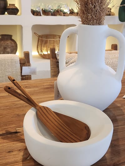 In a softly lit interior, likely a home decor store or a well-styled residence, a large white amphora-shaped vase filled with dried grass stands prominently on a wooden table. Beside it, a simple white bowl cradles two wooden spatulas, their handles engraved with a brand name that is partially obscured but appears to read "plum & oak". The background reveals shelves with decorative items, including rustic baskets and vases, adding to the overall natural and minimalist aesthetic. The scene is clean and uncluttered, suggesting a focus on texture and natural materials. The location is not specified in the image and cannot be determined.