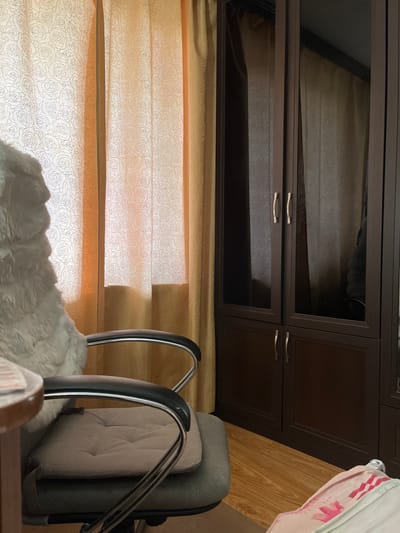 The image shows an indoor scene, likely a room. In the foreground, a grey office chair with a fluffy white seat cover is positioned. To the right of the chair is a dark brown wardrobe with mirrored doors on the upper section and solid doors on the lower section, featuring silver handles. Behind the chair and to the left, there are light yellow curtains with a subtle rose pattern. The floor is made of light brown wooden planks. The scene does not provide any specific visual cues to identify the location as Lytkarino, Russia. No people or actions are depicted.