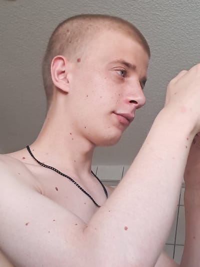 The image is a close-up, vertically oriented shot of a young person's head and upper body. The subject has a shaved head and is looking upwards and to the left, with their right arm raised. They are wearing a thin, black chain necklace. The background appears to be a textured wall, possibly in a bathroom, with a tiled surface visible further back. There are several moles visible on the subject's skin, including on their face, neck, and arm. The lighting suggests it is daytime, possibly indoors. The overall tone of the image is neutral, focusing on the subject's profile and skin details. The location is identified as Parchim, Germany.