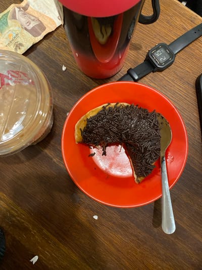 The image presents an overhead view of a dark brown wooden table, presumably in Pontianak, Indonesia. A bright orange circular plate centrally features a partially consumed crescent-shaped pancake, densely covered with dark brown chocolate sprinkles (mese). A silver spoon rests on the right side of the plate, partially embedded in the sprinkles. To the left of the plate, a clear plastic container with a domed lid contains a light brown substance. Behind the plate, a dark metallic or black insulated mug with a reflective surface and a red lid is visible. A crumpled 10,000 Indonesian Rupiah banknote is positioned to the left of the mug. To the right of the mug, a black Alexandre Christie digital wristwatch, displaying "2:15", lies on the table. A portion of a black object, likely a smartphone, is visible at the far right edge of the frame. Small white specks, consistent with food crumbs, are scattered on the tabletop.