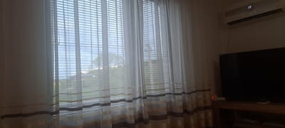 A sparsely lit interior scene, likely a room, is captured with a focus on a window covered by white, striped curtains and Venetian blinds. The view through the window suggests an exterior scene of green lawns or fields, possibly near a body of water, with a tree visible in the distance.
To the right of the window, an air conditioning unit is mounted high on the wall. Below it, a dark television screen dominates the space, placed on a wooden cabinet or stand that also holds some objects, possibly decorative or functional items.
The location is identified as Mineralni Bani, Bulgaria; however, no information about the exact street address is provided in the picture. There are no people visible, no actions taking place, and no interactions evident in the frame. The environment appears domestic, but the lack of details prevents a more specific characterization.