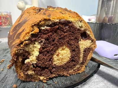 A cross-section of a marble cake is presented on a dark, possibly slate, serving plate. The cake has a distinct swirl pattern of chocolate and vanilla batter. The crust has a slightly browned and irregular appearance. The cake is positioned close to the viewer, dominating the frame. In the background, a blurred assortment of items is visible, including a jar of colorful candies, a white ceramic figurine, and a metal container. A silver knife partially rests on the plate alongside the cake. The surface under the plate appears to be a textured, light gray or white material. The location is Yanino-1, Russia.