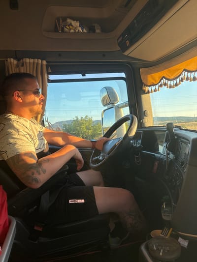 A man is driving a truck during the day, illuminated by the warm light of the setting sun. He is wearing sunglasses, a t-shirt with a speckled pattern, and black shorts. He has tattoos on his arms and legs. The truck's interior is visible, with the dashboard, steering wheel, and a gear shift. There are also curtains and decorative elements inside the cab. Outside the window, a rural landscape with trees and hills can be seen under a clear blue sky. The image seems to be taken from the passenger seat, looking towards the driver. The location is the city of Aetokoryfi, Greece.