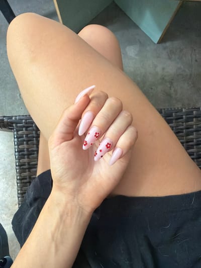 A close-up, high-angle shot shows a person's legs and hand, with the person seated in a wicker chair. The person's legs are bare and tanned, and they are wearing black shorts. The hand is held up, displaying light pink stiletto nails adorned with small red floral designs. The background consists of a concrete floor and a turquoise wall. The scene appears to be indoors, possibly in a salon or home environment. There is no specific information in the image to determine its location within Prosena, Bulgaria.
