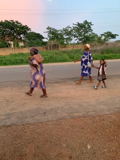 The image shows three individuals walking along a paved road in Gbeleko, Nigeria, under the dusk sky. Two adults, presumably women, are seen wearing long dresses. One has a satchel. The third individual, a young girl, is walking hand-in-hand with one of the women. Trees and greenery line the road. Power lines are visible against the sky. The edge of the road is a mix of gravel and dirt.
