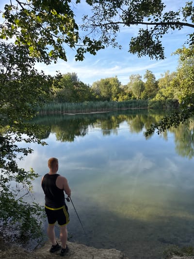 The image shows a man fishing on the shore of a lake or pond. He is standing on a dirt embankment, facing the water. He is wearing a black tank top and black shorts with yellow stripes. He is holding a fishing rod. The water is calm and reflects the trees and sky. In the background, there is a line of reeds and more trees. The sky is blue with some white clouds. The image appears to have been taken on a sunny day. The location is likely the Baggersee in Ludwigshafen am Rhein, Germany, a popular recreational area.
