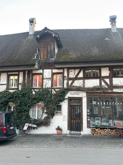 The image displays the facade of a traditional half-timbered building located in Eglisau, Switzerland, under a bright, overcast sky. The two-story structure features white stucco walls with dark brown exposed wooden beams forming a rectangular grid pattern. A dark gray tiled roof, partially covered in moss, has two brick chimneys and a prominent wooden dormer window with vertical slats.
On the ground floor, from left to right, a black Volkswagen California van is parked on a cobbled street, adjacent to a metal A-frame sign displaying a red "No Parking" symbol and a white bicycle wheel. Extensive green ivy and bare vines cover a significant portion of the building's left side, intertwining around a ground-floor window and extending across the facade towards a dark wooden entrance door. Above this door, a white sign reads "Landjägeramt." To the right of the door, a small terra-cotta pot contains a plant. Further right, a large storefront window displays various artworks, identifying the establishment as "Malatelier IRI-ART." Below this window, a neatly stacked pile of firewood is visible, alongside a small red metal outdoor chair.
The upper floor features multiple windows; two centrally located windows exhibit an internal orange glow, possibly from lighting or curtains, contrasting with the other windows which show ambient light. No individuals are present in the scene. The street in the foreground is asphalt, featuring a single circular manhole cover.
