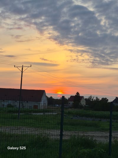 The image depicts a sunset over a rural landscape, possibly in Pątnów, Poland, given the "Galaxy S25" watermark which may indicate the origin of the photo. The sky is a canvas of warm colors, transitioning from a bright orange and yellow near the horizon to softer pastel hues and gray clouds higher up. The sun, a vibrant red-orange disc, is partially obscured by clouds and appears low on the horizon, indicating either sunrise or sunset.
In the foreground, a dark wire fence runs horizontally across the frame, partially obscuring a grassy area with some sparse vegetation. Beyond the fence, a long, single-story building with a reddish-brown roof is visible. Its facade appears weathered and light-colored. To the left of this building, a utility pole with insulators and wires stands prominently.
Further in the background, a few houses with pitched roofs and trees are scattered across the landscape. The overall scene conveys a sense of peaceful rural twilight. There are no people or animals visible in the image. The image is taken from a slightly elevated perspective, looking towards the horizon.