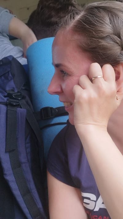 This is a close-up, waist-up, outdoor portrait of a young woman with fair skin and braided brown hair. She is looking to her left, with a slight smile and her right hand is touching her face near her ear. She is wearing a dark blue t-shirt with white text that is partially visible, appearing to say "SA". She has a thin gold ring on her right ring finger. Behind her, a dark blue backpack is visible, along with a bright blue rolled-up mat. The background suggests a casual, outdoor setting, possibly a park or campsite. The lighting is bright, indicating it is daytime, and there are no visible weather cues. The overall mood appears relaxed and candid. The image was taken in Osel'ki, Russia.