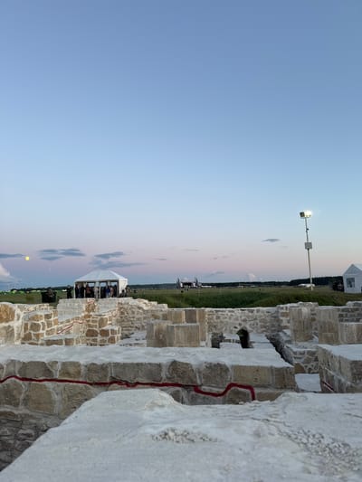 The image shows the archaeological ruins of the Great Bulgar city, located in Bolgar, Russia. In the foreground, ancient stone walls with red markings are visible. In the mid-ground, a white tent is set up, with several people standing around it. Further in the background, there are green fields, tents, and a light post with a spotlight. The sky is a gradient of pale blue and pink, indicating either dawn or dusk. A full moon is visible in the upper left corner of the sky.