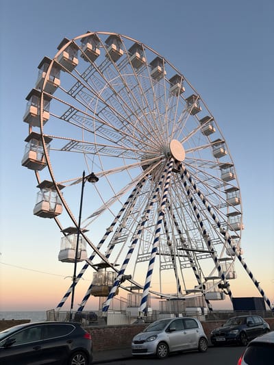 The image displays a large white Ferris wheel under a clear sky, captured from a low angle. The Ferris wheel, the primary subject, features numerous passenger gondolas and has several of its structural spokes wrapped in a distinctive blue and white striped pattern. A circular, textured hub is visible at the center of the wheel, and a light fixture is attached to one of its support beams.
The sky exhibits a gradient, transitioning from a light blue at the top to soft hues of orange, yellow, and pink towards the bottom right, suggesting either dawn or dusk.
To the right of the Ferris wheel, several cars are parked on a paved area, likely a street or promenade. A silver Fiat Punto with the UK license plate "AY63 HFM" is prominently visible, as is a dark blue BMW 1 Series with the UK license plate "NV12 LNN". Parts of other vehicles, including a dark-colored car in the foreground and a red car in the upper right corner, are also present. Beyond the parked cars, a low barrier or wall separates the paved area from the calm sea, which extends to the horizon. No people are explicitly visible, and no specific activity beyond the presence of the Ferris wheel and parked cars is taking place. The scene suggests a quiet moment at a coastal location, consistent with East Suffolk, United Kingdom.
