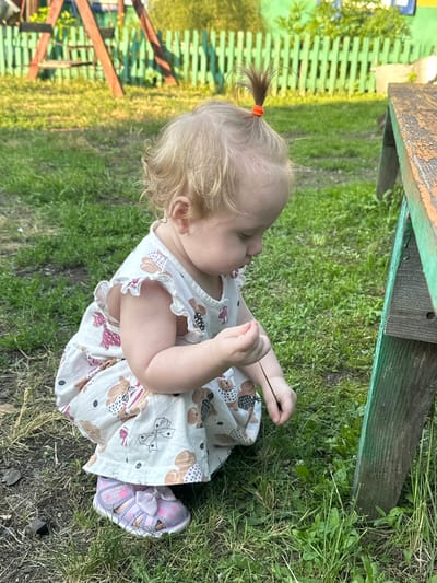 The image depicts a young child, likely a toddler, crouching on the grass in what appears to be a backyard. The child has light blonde hair pulled into a small ponytail with an orange hair tie. They are wearing a light-colored dress with a pattern of brown bears and pink bows. On their feet are lavender sandals with pink bows. The child is intently looking down at their hands, holding a thin twig. To the right of the child is a weathered wooden object, possibly a bench or part of a structure, painted green on the side and with orange accents on the top edge. In the background, a green wooden fence and a wooden swing set are visible. The ground is covered in green grass and some dry earth. The lighting suggests it is daytime, likely late afternoon, with the sun casting shadows. This scene is situated in the city of Novoyeniseyka, Russia.